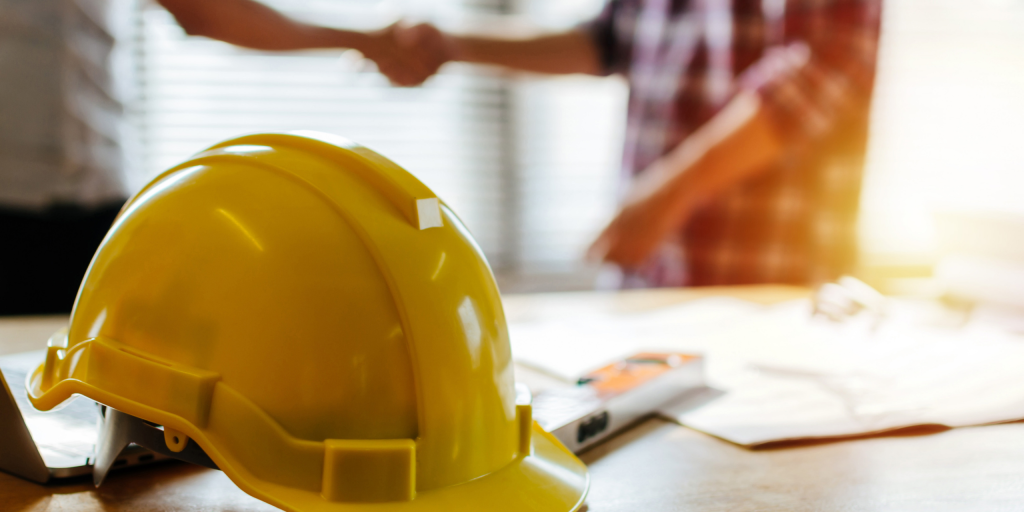 There is a yellow hard hat on the left on desk with two people blurred out in the background making a deal about excavation services.